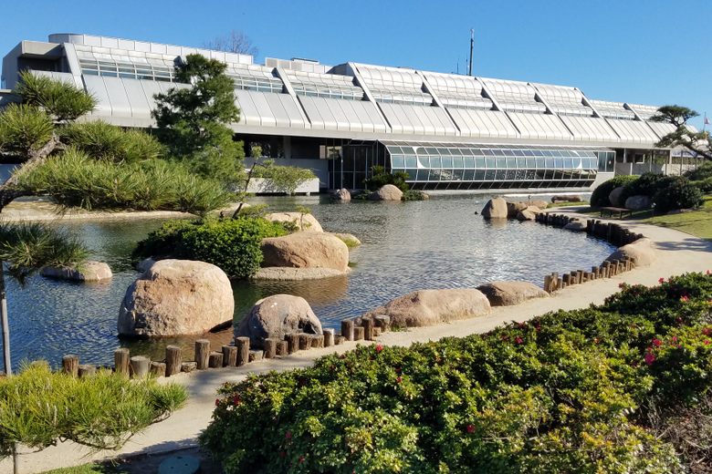 A large, modern glass and metal building overlooks a Japanese garden with a pond, large rocks, and manicured shrubs.