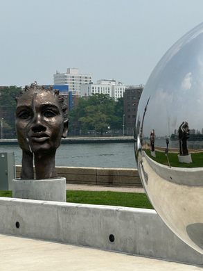 A split bronze sculpture of a Black woman with a river behind and a mirrored orb on the right