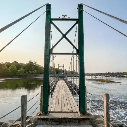 A narrow wooden footbridge with green steel towers crossing a tidal river at sunset