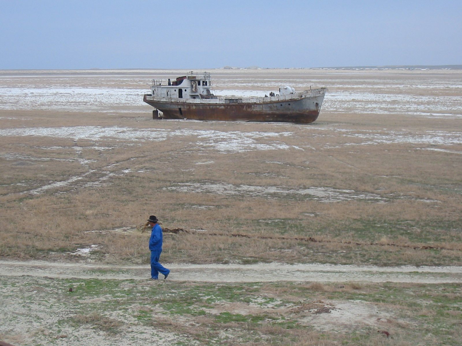 A man walks past an orphaned ship rusting away in the desert.