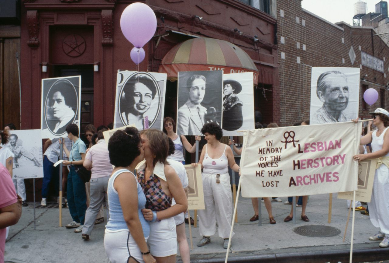 Women kiss, celebrate, and hold up a banner for the Lesbian Herstory Archives in front of the Duchess Bar, then a popular lesbian bar in Greenwich Village, at Pride in 1982.