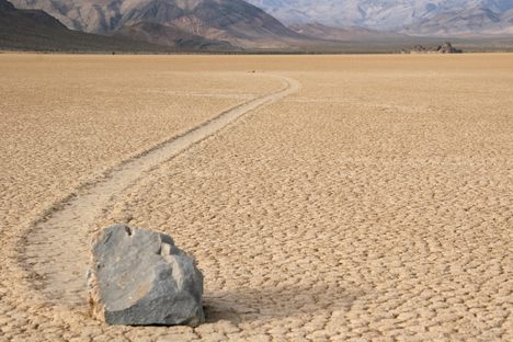 A medium gray rock on the desert floor, with a trail in the dirt behind it showing how the rock has moved.