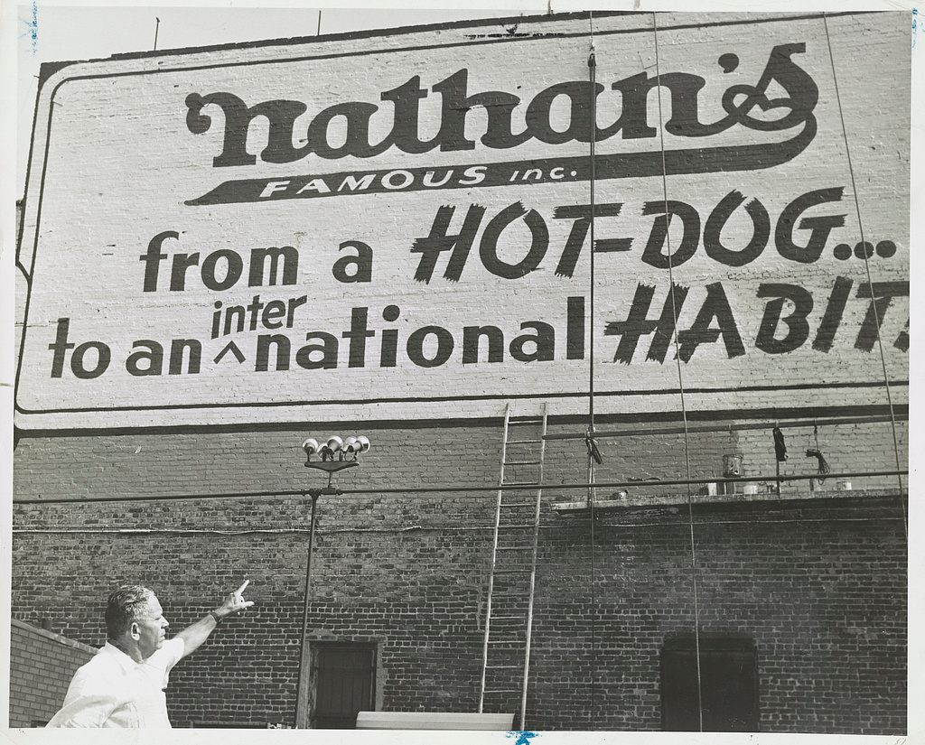 Photograph showing Nathan Handwerker pointing to restaurant sign painted on outside wall.