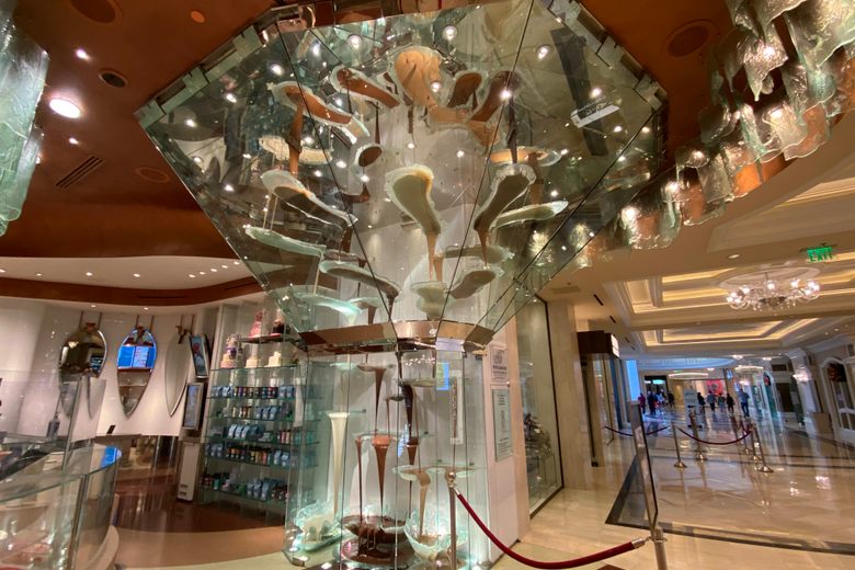 A tall glass-encased fountain cascades dark, milk, and white chocolate down tiered glass basins in a shop.