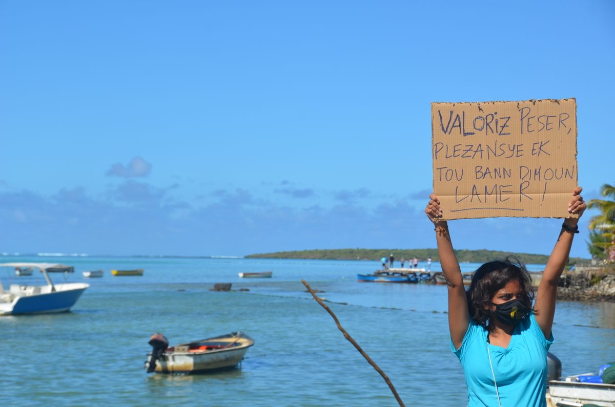Activist Shaama Sandooyea, one of hundreds of volunteers at the People's Factory Zone, holds up a sign on the Mahebourg waterfront that translates loosely as "Value fishermen, boaters, and people of the sea."