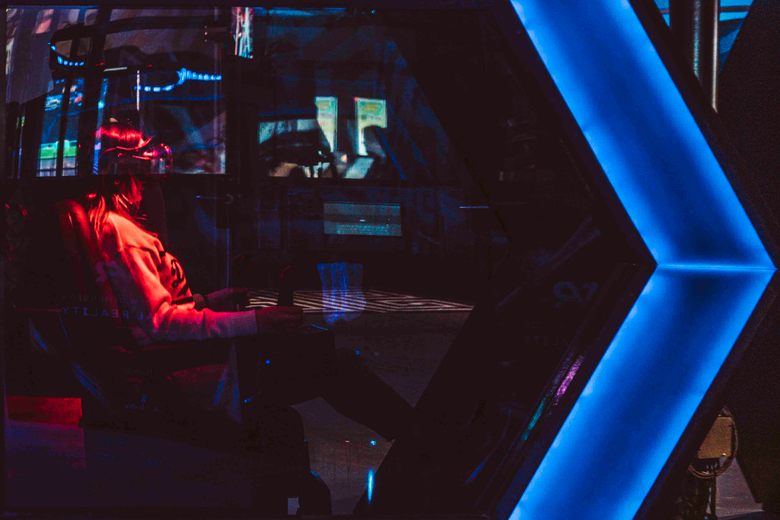 A person wearing a VR headset sits in a dark gaming pod illuminated by red and blue neon lights.