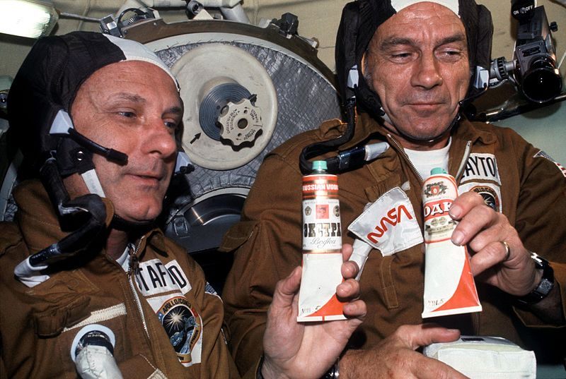 July 1975: Astronauts Thomas P. Stafford (left) and Donald K. "Deke" Slayton hold containers of Soviet space food in the Soyuz Orbital Module during the joint U.S.-USSR Apollo-Soyuz Test Project docking in Earth orbit mission. The containers hold borscht (beet soup) over which vodka labels have been pasted. 