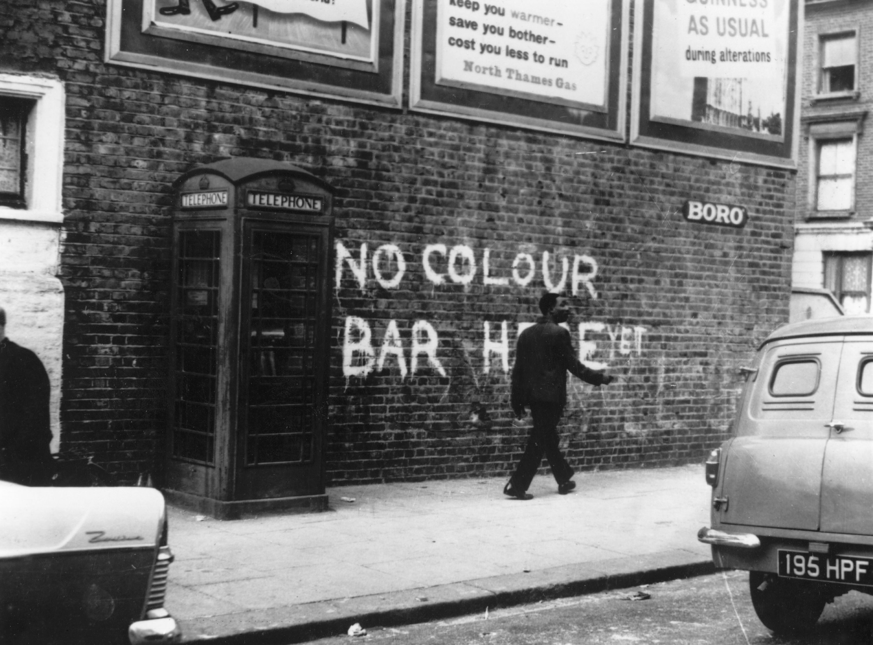 A Black man walks down Notting Hill Street and past racist graffiti, circa 1959.