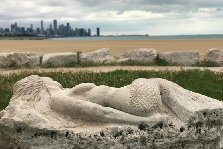 A stone sculpture of a reclining mermaid overlooks a sandy beach and the Chicago skyline across the water.