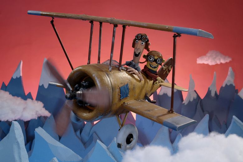 Two claymation characters wearing goggles fly a small yellow biplane over jagged blue mountains and cotton clouds.