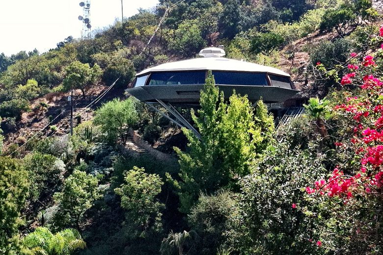 The Chemosphere, a white octagonal saucer-shaped house, perched on a concrete pedestal above a lush, wooded hillside.