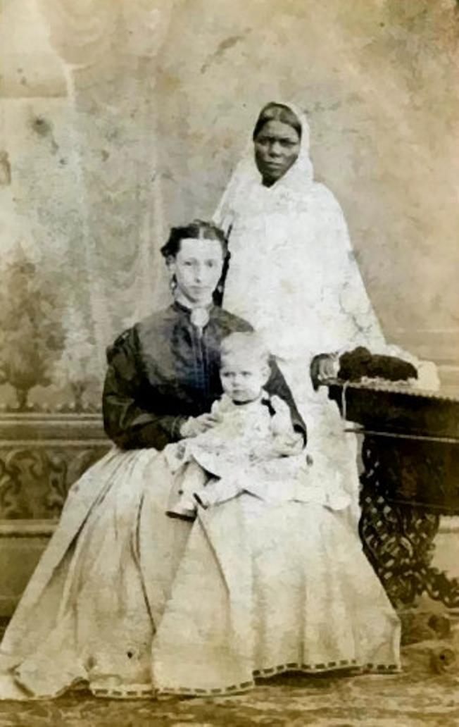 An ayah stands behind a woman and her child, location unknown, in this image from the 1860s.