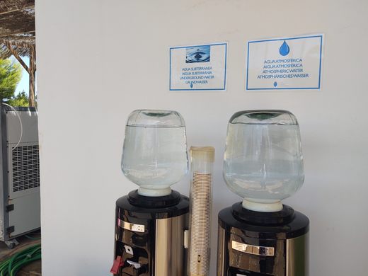 Two water jugs in water dispensers in front of a white walls with signs