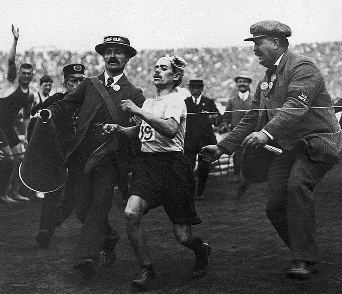 Dorando Pietri is helped across the finish line while holding a cork in his hand.