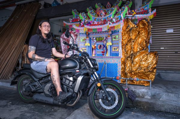 Metal guitarist and paper effigy artisan Koh Eng Keat sits on his motorcycle in front of a seven-foot-tall paper mansion. Keat runs 358 Custom Effigies Workshop with his father Koh Ah Bah where building elaborate papier-mâché effigies like this are just part of a normal workday.